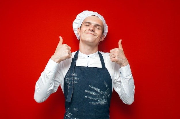 Gelukkige kok toont zoals met handen op rode geïsoleerde achtergrond kerel in chef-kokuniform glimlacht