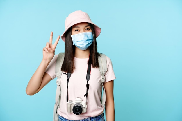 Gelukkige Koreaanse vrouw in medische gezichtsmasker fotocamera met vredesvsigns die reizen tijdens covid pa...