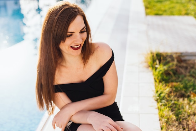 Gelukkige langharige jonge vrouw in een zwarte jurk zit bij het blauwe zwembad, lachend in de zon