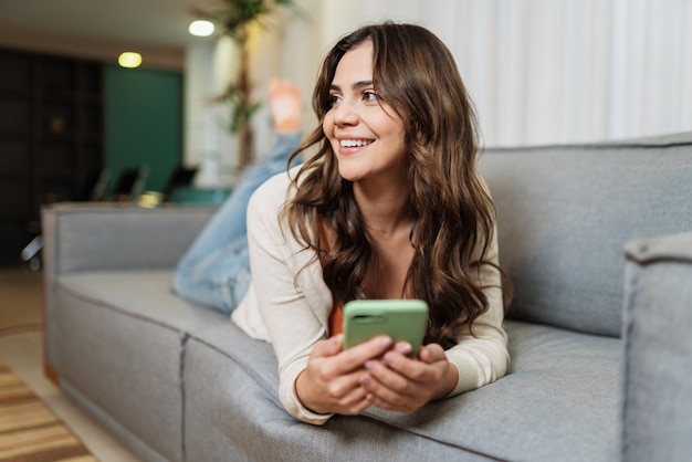 Gelukkige Latijnse dame ontspant alleen thuis op kamer pose en deelt goed nieuws op sociale media via mobiele telefoon. Glimlachende vrouw geniet van weekendbestelling goederen online in de telefoon-app.