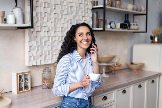 Gelukkige latijnse vrouw die koffie drinkt en een telefoontje heeft dat in het keukeninterieur staat en glimlacht