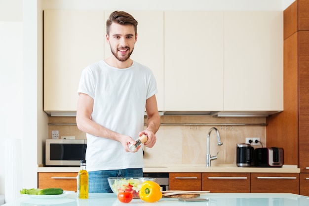 Gelukkige man die kruiden in salade toevoegt in de keuken en naar de voorkant kijkt