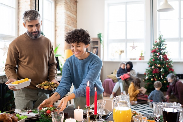 Gelukkige man en vrouw die zelfgemaakte salade, gebakken aardappelen, drankjes en ander voedsel op een feestelijke tafel zetten voor het familiekerstdiner