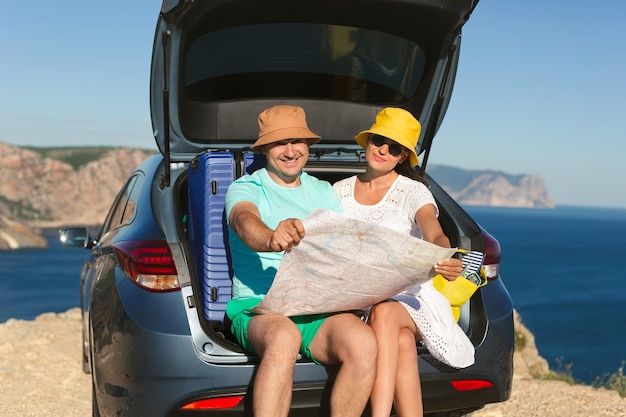 Gelukkige man en vrouw zitten in de kofferbak van een auto aan zee met een wegenkaart in hun handen