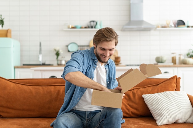 Gelukkige man klant uitpakken kartonnen doos ontvangt open postbezorgingspakket. Glimlachende jonge tevreden shopper unboxing pakket uitpakken verzending bestelling productaankoop thuis op de bank zitten