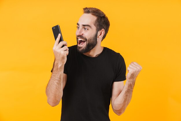 gelukkige man met een basic zwart t-shirt die zich verheugt en praat op smartphone geïsoleerd over gele muur