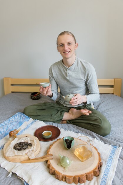 Gelukkige man zittend op het bed, doet de theeceremonie alleen, blijf thuis en wees veilig tijdens de carantine. Chinese thee drinken