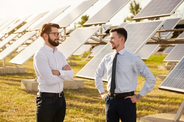 Gelukkige mannelijke zakenpartners bespreken energieontwikkelingsproject terwijl ze in de buurt van fotovoltaïsche panelen op groene zonneboerderij staan