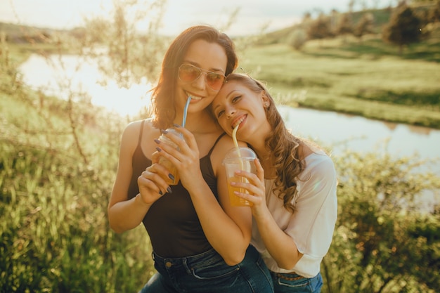 Gelukkige meisjes die pret hebben, die cocktail plastic kop met een stro in zonnebril drinken die de camera bekijken, bij zonsondergang, positieve gelaatsuitdrukking, openlucht