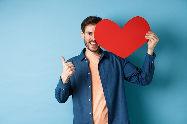 Gelukkige mens die valentijnskaartenhart en thumbs-up gebaar tonen, die zich over blauwe achtergrond bevinden.