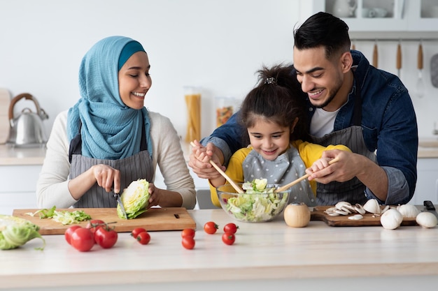 Gelukkige Midden-Oosterse familie met dochtertje koken salade In de keuken samen, liefdevolle Arabische vader helpen zijn kind om groenten te mengen In de kom, genieten van het bereiden van voedsel thuis, close-up