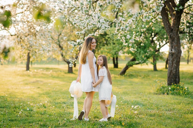 Gelukkige moeder en haar dochter in witte jurken wandelen in de lentetuin bij zonsondergang