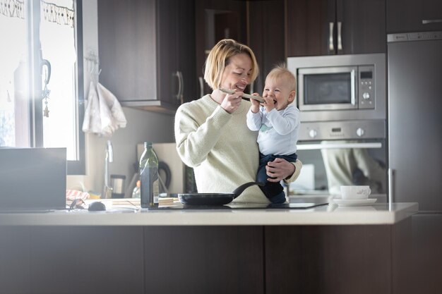 Gelukkige moeder en kleine baby koken en proeven een gezond diner in de huishoudelijke keuken