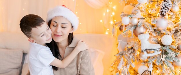 Gelukkige moeder en zoon met een kerstmanhoed knuffelen op bed in een versierde slaapkamer op kerstavond in