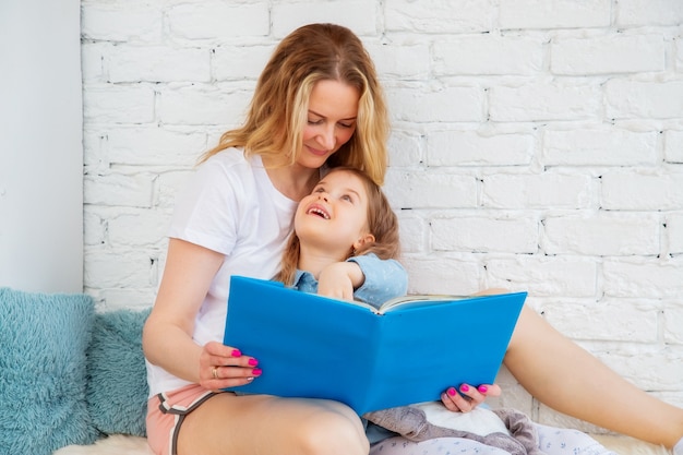 Gelukkige moeder leest een vrolijk sprookje in een boek voor aan haar dochter en glimlacht terwijl ze in de woonkamer zit