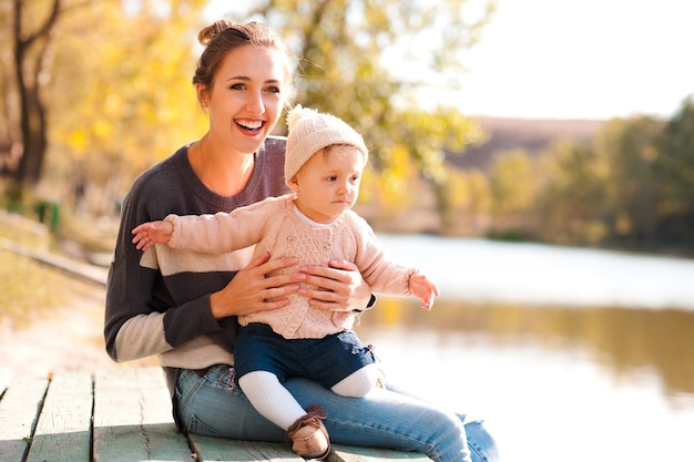 Gelukkige moeder met babymeisje van 1 jaar oud met gebreide truien op een houten pier in het herfstpark Wegkijken Geluk