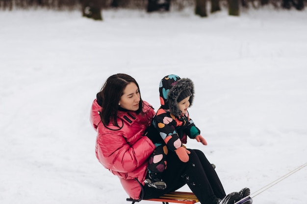 Gelukkige moeder met babymeisje zittend op een slee en slee naar beneden op sneeuw vanaf de heuvel