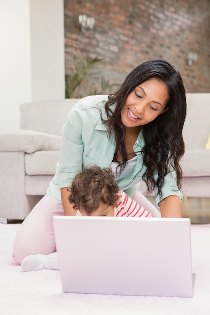 Gelukkige moeder met haar baby die laptop op het tapijt in woonkamer met behulp van