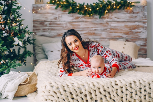 Gelukkige moeder met haar dochtertje in vakantie kleding met gedrukte herten en sneeuwvlokken plezier op het bed in gezellige kamer met een kerstboom en kerstverlichting. Nieuwjaar en Kerstmis concept.