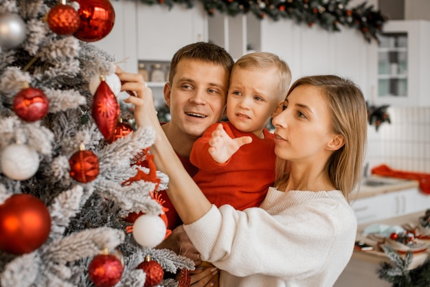 Gelukkige moeder, vader en zoontje versieren samen kerstboom thuis, kleine jongen huilt en wil speelgoedbal in zijn arm nemen