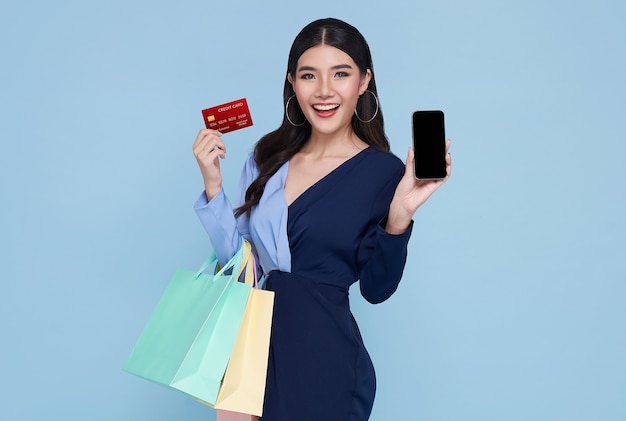 Gelukkige mooie Aziatische vrouwen die creditcard en smartphone tonen en boodschappentassen houden die op blauwe achtergrond worden geïsoleerd.