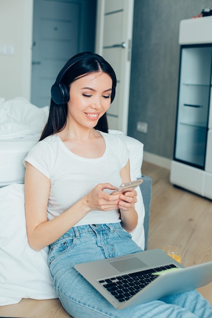 Gelukkige mooie dame die een koptelefoon draagt terwijl ze bij het bed zit met een laptop op haar schoot en een smartphone gebruikt