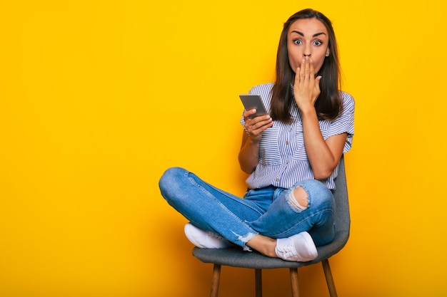 Gelukkige mooie stijlvolle vrouw zit op de stoel en gebruikt haar smartphone terwijl ze geïsoleerd is op gele achtergrond