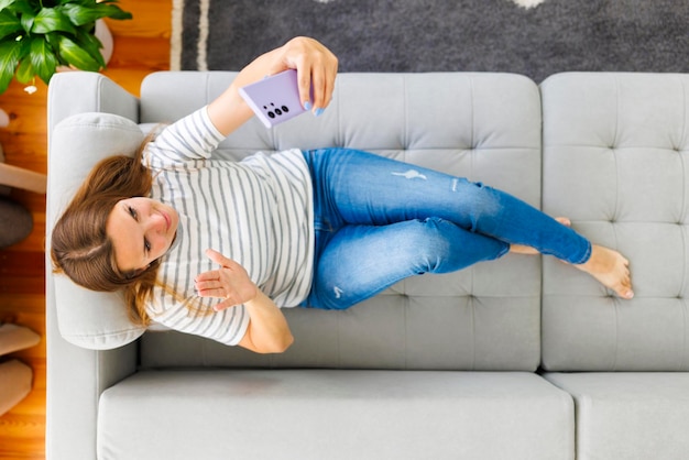 Gelukkige mooie vrouw die aan de telefoon praat terwijl ze thuis op de bank ligt.