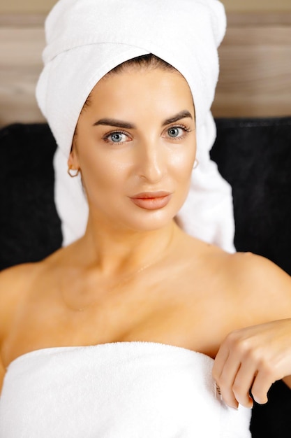 Gelukkige mooie vrouw die badjas en witte handdoek draagt met perfecte teint wat betreft gezicht kijkend naar camera zittend in kamer Reclame voor huidverzorging spa wellness-concept Close-up portret