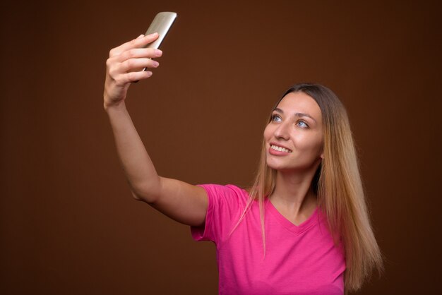 Gelukkige mooie vrouw die mobiele telefoon met behulp van om selfie tegen bruine muur te nemen