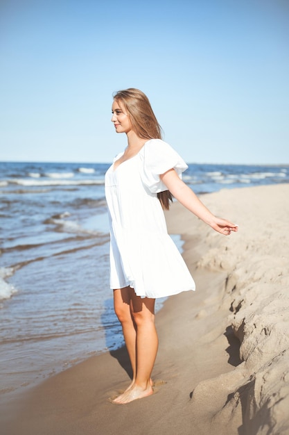 Gelukkige, mooie vrouw op het strand van de oceaan, staande in een witte zomerjurk, open armen.