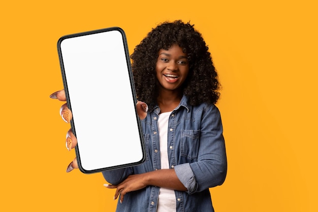 Gelukkige mooie zwarte vrouw op gele achtergrond die smartphone toont