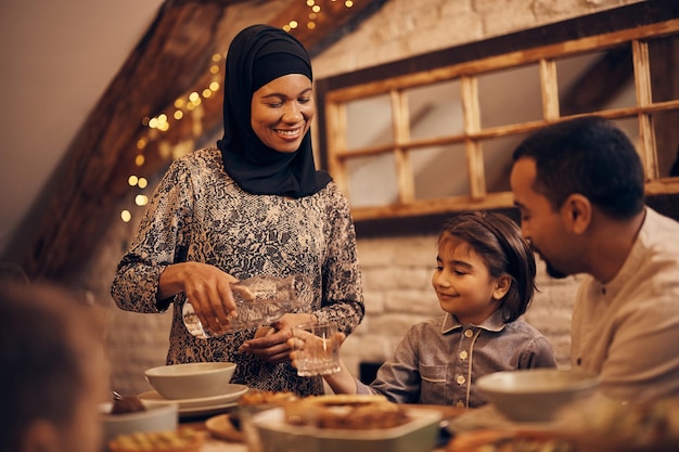 Gelukkige moslimvrouw die water in het glas van haar dochter giet tijdens een gezinsdiner aan de eettafel op Ramadan