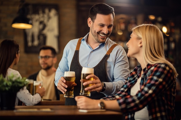 Gelukkige ober die communiceert met vrouwelijke gast terwijl ze bier serveert aan haar tafel in een bar