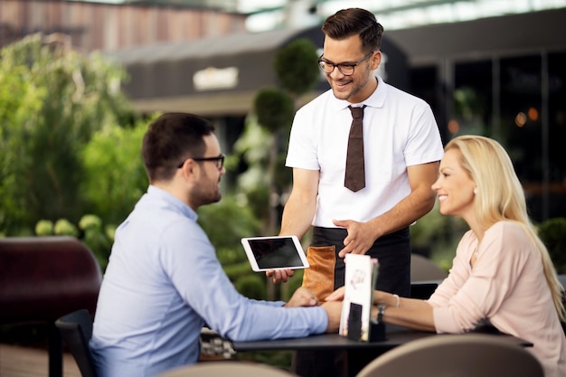 Gelukkige ober die digitale tablet gebruikt en het menu aan zijn gasten in een café laat zien