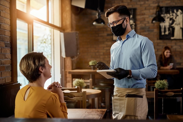 Foto gelukkige ober die met vrouwelijke klant praat terwijl hij een beschermend gezichtsmasker en handschoenen draagt in een café