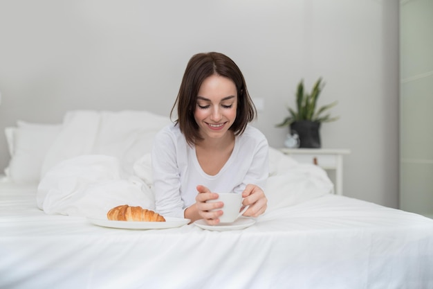 Gelukkige ontspannen jonge vrouw die koffie drinkt en gebak eet in bed