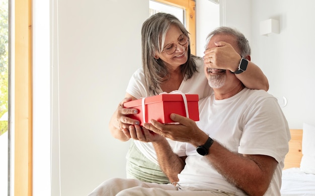 Gelukkige oude europese vrouw sluit de ogen voor haar man geeft een geschenkdoos in het interieur van de woonkamer