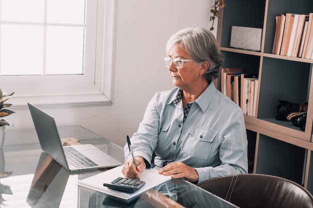 Gelukkige oude kaukasische zakenvrouw glimlachend online werken kijken naar webinar podcast op laptop en leren onderwijs cursus conferentiegesprekken maken aantekeningen zitten op het werk bureau, e-learning concept