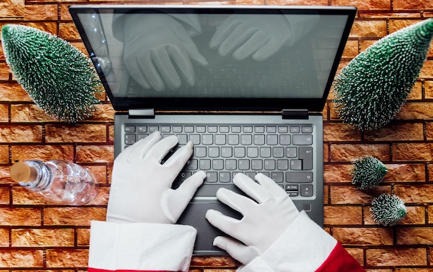 Gelukkige oude kerstman met hoed die een geschenkdoos vasthoudt met behulp van een laptopcomputer die laat op een vrolijke kerstavond aan de thuistafel van de workshop zit. E-commerce website kerstmis vakantie online winkelen e-commerce verkoop