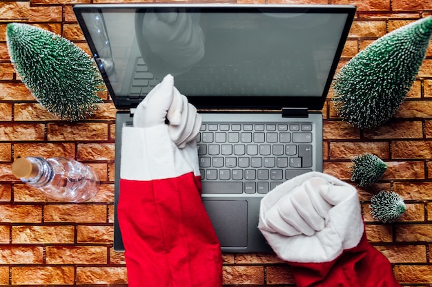 Gelukkige oude kerstman met hoed die een geschenkdoos vasthoudt met behulp van een laptopcomputer die laat op een vrolijke kerstavond aan de thuistafel van de workshop zit. E-commerce website kerstmis vakantie online winkelen e-commerce verkoop