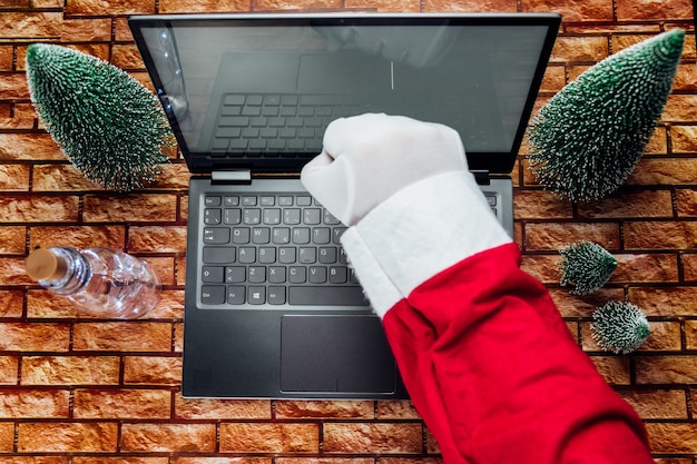 Gelukkige oude kerstman met hoed die een geschenkdoos vasthoudt met behulp van een laptopcomputer die laat op een vrolijke kerstavond aan de thuistafel van de workshop zit. e-commerce website kerstmis vakantie online winkelen e-commerce verkoop