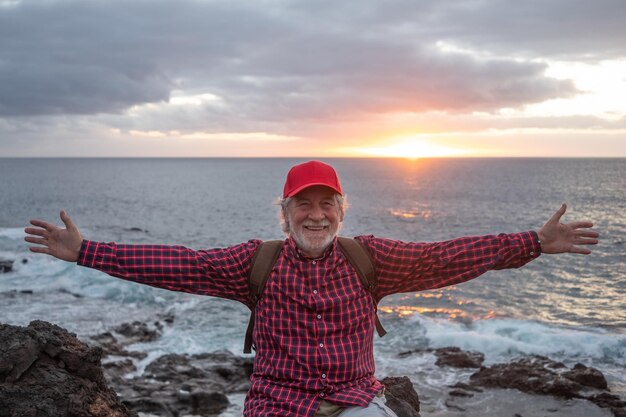 Gelukkige oude senior man met rode dop buiten op zee zitten bij zonsondergang met uitgestrekte armen knappe oudere man genieten van vrijheid en vakantie