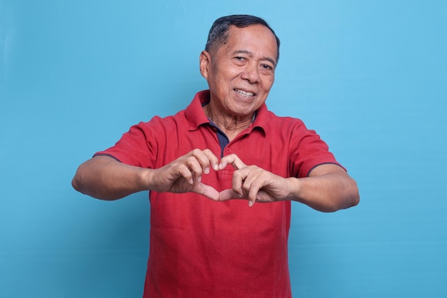 Gelukkige oudere man met casual rood t-shirt over geïsoleerde blauwe achtergrond glimlachend en hart tonend