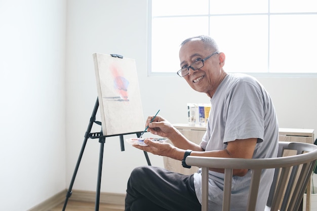 Gelukkige oudere man met een bril die op een stoel zit en naar de camera kijkt terwijl hij op doek schildert