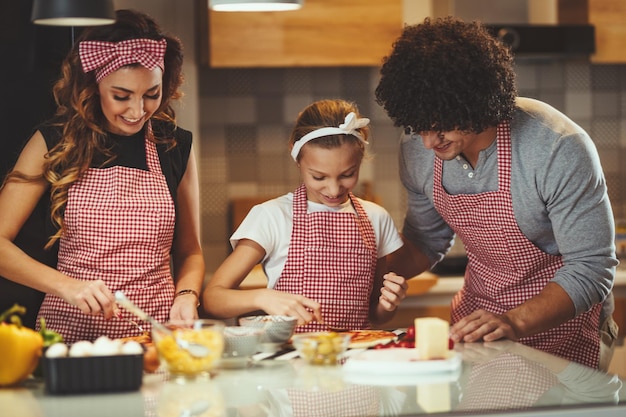 Gelukkige ouders en hun dochter bereiden samen een maaltijd in de keuken terwijl het meisje ketchup op het pizzadeeg doet.