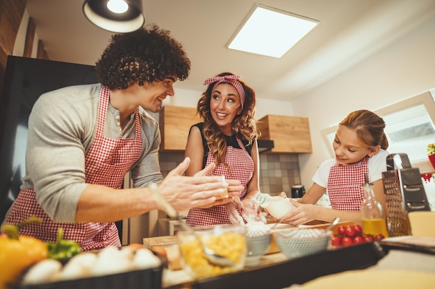 Gelukkige ouders en hun dochter bereiden samen koekjes in de keuken. Klein meisje helpt haar ouders de olie in de schotel met deeg te gieten.