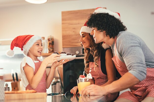 Gelukkige ouders en hun dochter met kerstmuts bereiden samen een maaltijd in de keuken. Klein meisje houdt bord met muffin vast dat haar moeder en vader aanbiedt.