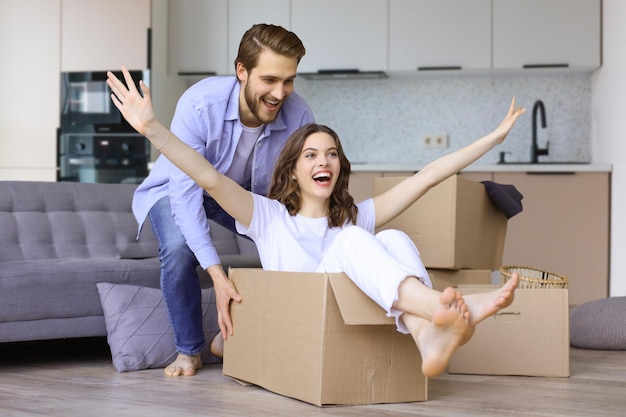 Gelukkige paar heeft plezier met kartonnen dozen in nieuw huis op de dag van de verhuizing vrouw rijden zittend in kartonnen doos terwijl man duwen