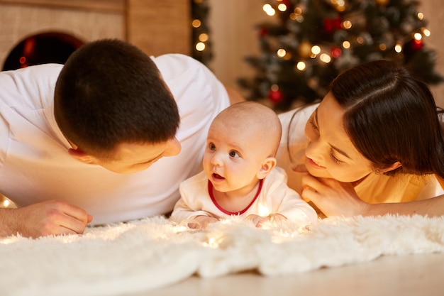 Gelukkige paar met baby liggend op de vloer op wit zacht tapijt, ouders kijken naar hun baby met grote liefde, binnenshuis poseren met kerstboom op de achtergrond, gelukkig nieuwjaar.